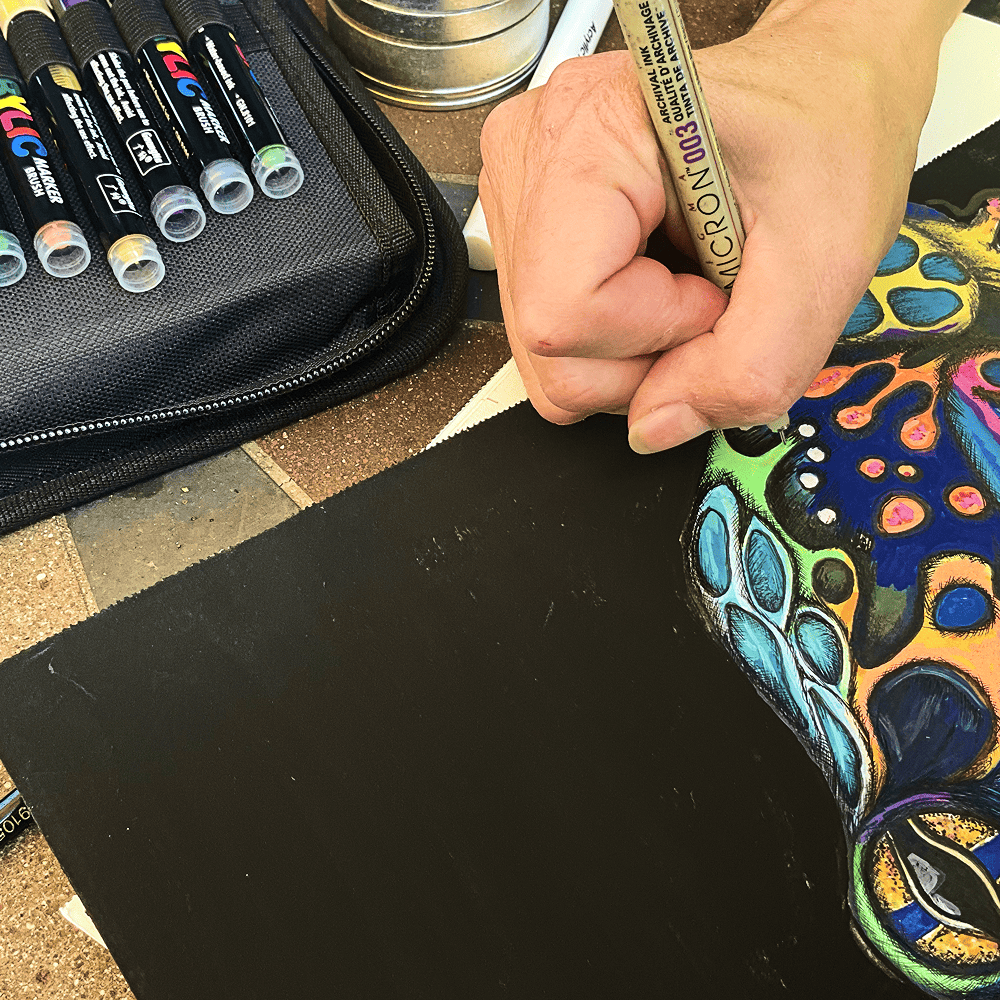 The Psychedelic Frog being drawn on black paper with a fine-point pen, acrylic markers alongside