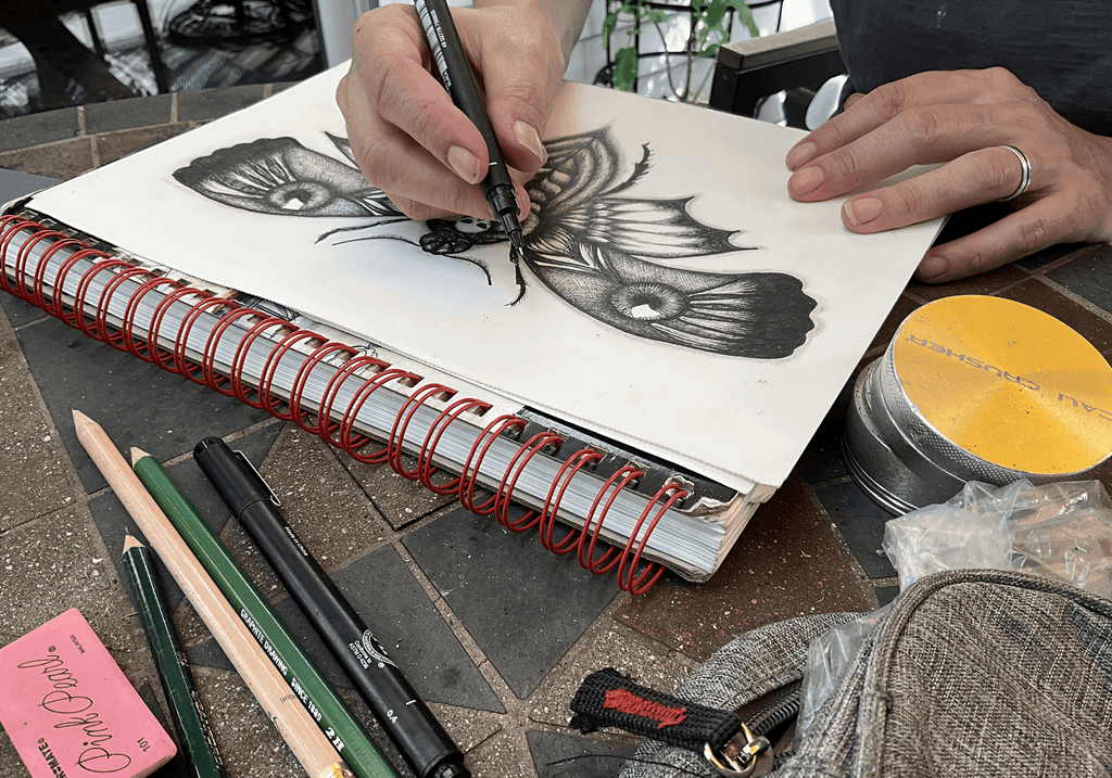 Both hands inking the moth's wings in a spiral-bound sketchbook