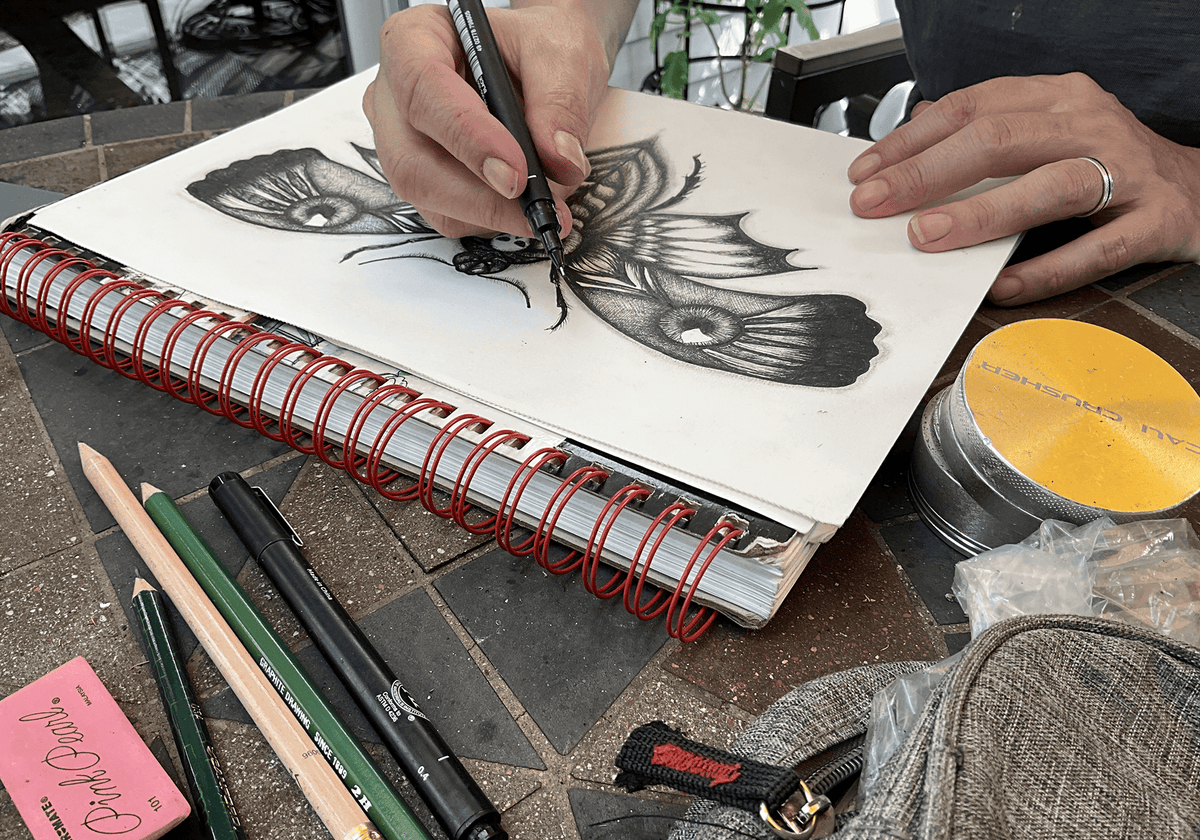 Both hands inking the moth's wings in a spiral-bound sketchbook